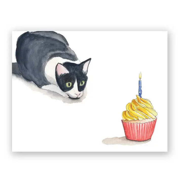 White greeting card featuring a watercolor illustration of a black and white cat poised to pounce on a cupcake with a single blue birthday candle in it
