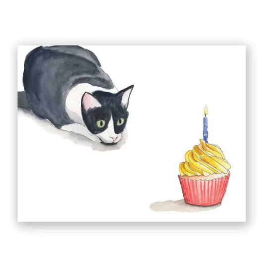 White greeting card featuring a watercolor illustration of a black and white cat poised to pounce on a cupcake with a single blue birthday candle in it