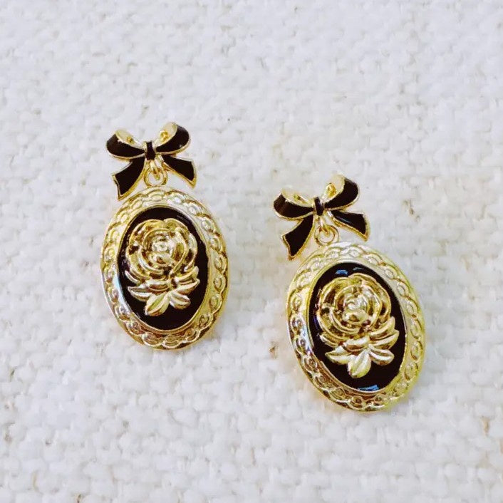 Pair of black and gold earrings shaped like framed roses hanging from bow-shaped studs