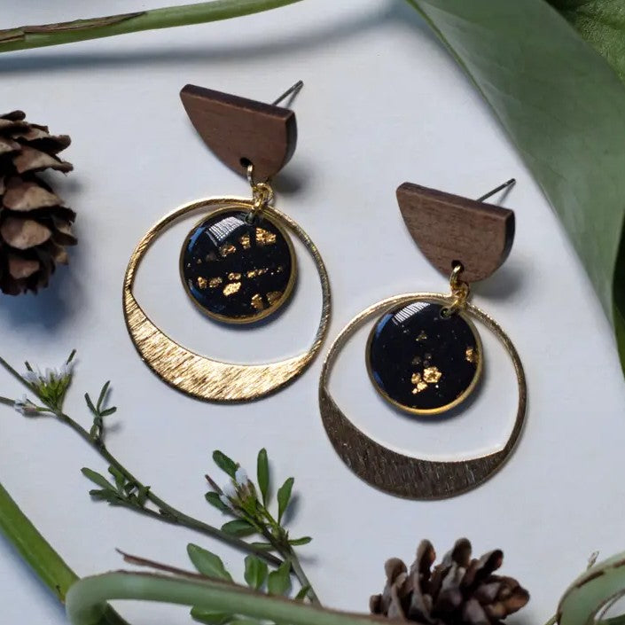 A pair of geometric drop earrings. The studs are wooden half circles, from which gold-flecked black pendants hang inside larger gold circles