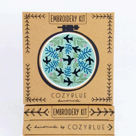 An embroidery kit featuring several black birds flying, surrounded by pink, blue and green foliage over a light green background.