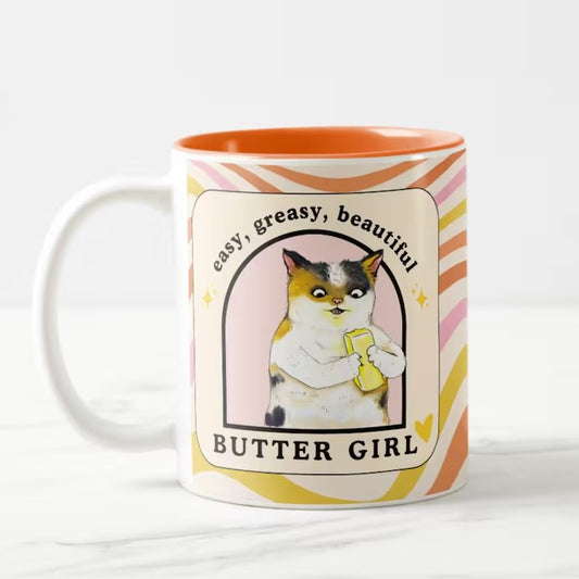 Mug with pink, orange and yellow swirls and an illustration of a cat eating a stick of butter with the caption "Easy, greasy, beautiful, butter girl"