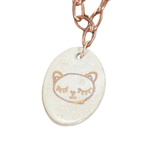 A close-up of a pendant on a copper chain. The pendant is a ceramic white oval with a gold cat face painted on it