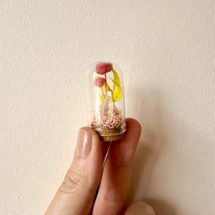 Person holding a tiny glass terrarium with pink flowers, moss and a small crystal inside