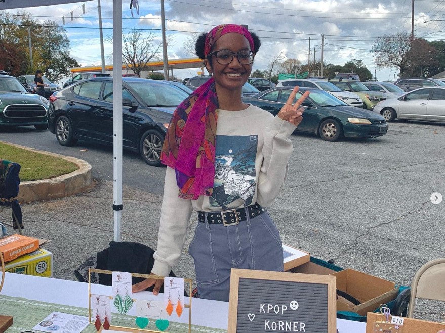 Fari of Love Nana Co. standing at a craft fair booth selling earrings and holding up a peace sign.