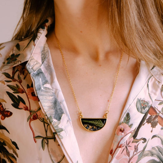 Gold plated necklace with a semicircle pendant featuring a small pressed fern frond and blue opal against a black resin background