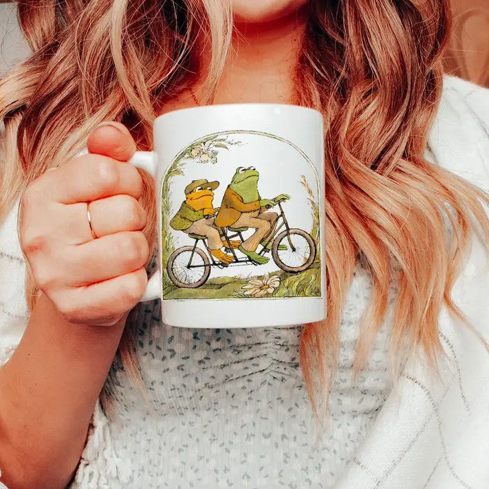 Person holding a white mug with an illustration of Frog and Toad riding a bicycle on it