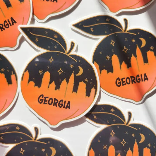 Small vinyl sticker shaped like a peach, depicting a black and orange Atlanta skyline