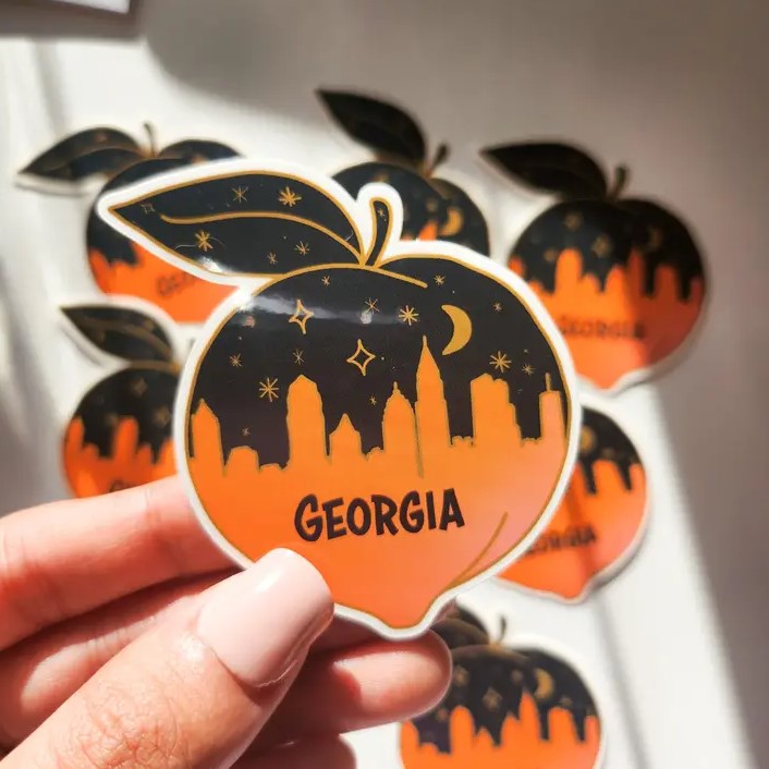 Small vinyl sticker shaped like a peach, depicting a black and orange Atlanta skyline