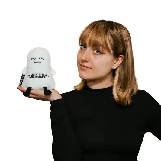 Person holding a white plush toy in the shape of a ghost with a blank expression and embroidered text that reads "Livin' the nightmare"