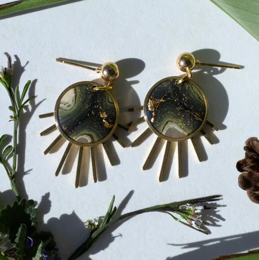 A pair of gold-tone drop earrings with circular agate-like stone pendants featuring green, black, and white marbled bands, with radiating gold spikes below. The earrings are styled on a white surface with natural botanical props.