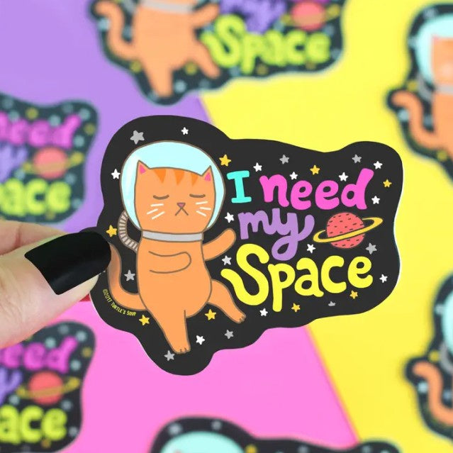 Sticker of an orange cartoon cat wearing a space helmet and drifting through the stars with the caption "I need my space"