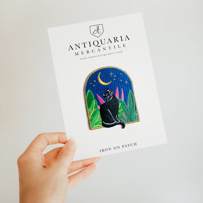 A hand holding a patch featuring a cat looking at the night sky with moon and stars surrounded by plants