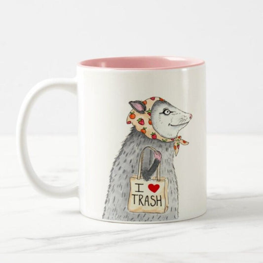 Mug with a pink interior and white exterior featuring a design of a possum in a strawberry-printed bandana carrying a tote bag that says "I <3 trash"