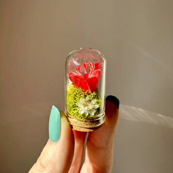 Person holding a tiny glass terrarium with a red flower and moss inside
