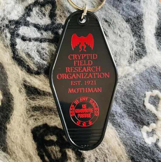 Black keychain with red lettering that reads "Cryptid field research organization est. 1921 Mothman"