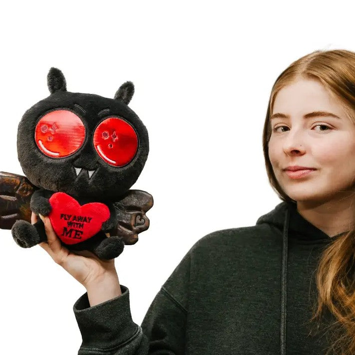 Person holding black plush toy shaped like Mothman holding a heart that reads "Fly away with me"