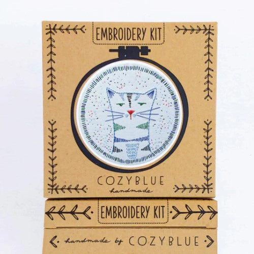An embroidery kit featuring a cat with a deadpan expression stitched in blue and green colors.