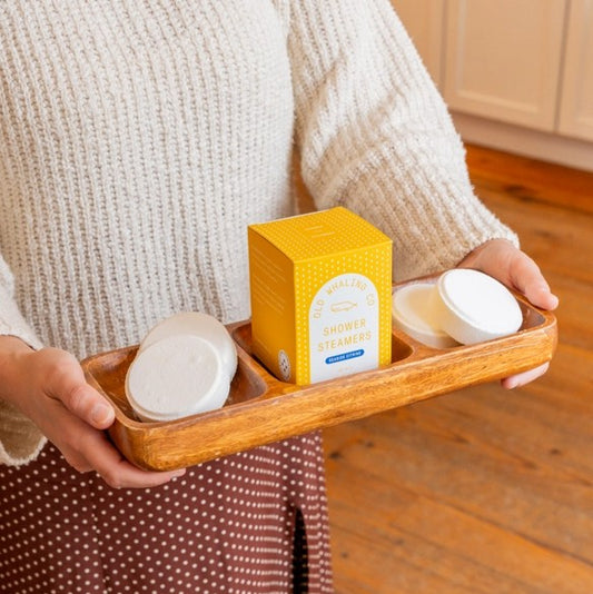 Person holding a wooden tray with four shower steamers and a bright yellow box on it