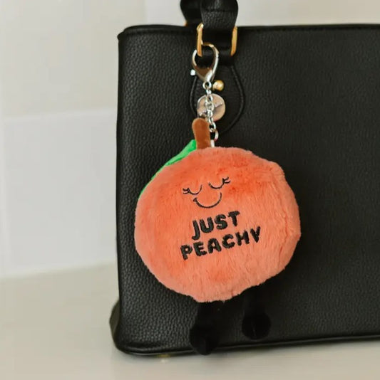 Peach plushie keychain with a peaceful smiling face and the words "Just Peachy" stitched on it in black thread