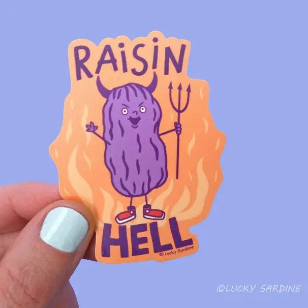 Sticker of an evil smiling raisin with devil horns on a fiery background with the text "Raisin Hell"