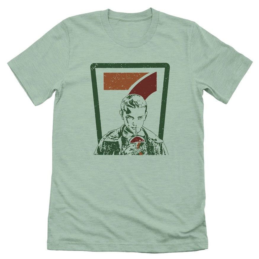 Light green t-shirt with the 7-11 logo behind Eleven from Stranger Things sipping a large drink