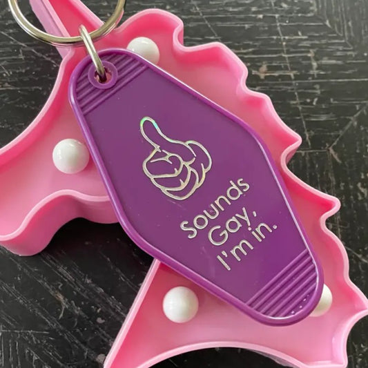 Purple keychain with a silver thumbs up and lettering that reads "Sounds gay, I'm in."