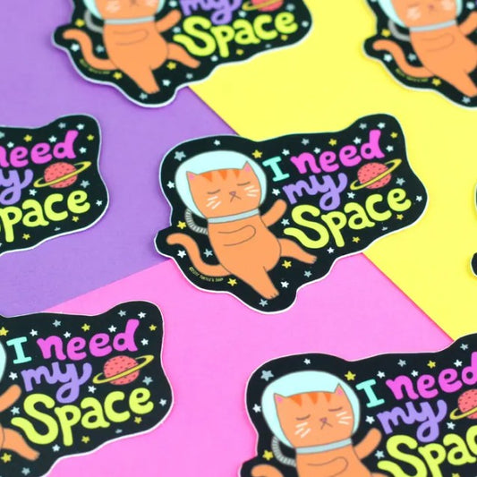 Sticker of an orange cartoon cat wearing a space helmet and drifting through the stars with the caption "I need my space"