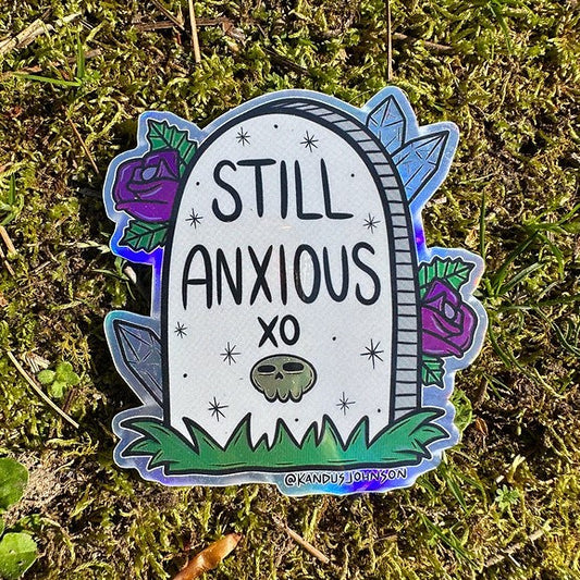 A holographic sticker featuring a headstone inscribed with a cartoon skull and the words "STILL ANXIOUS XO" in a handwritten font and surrounded by purple roses and gemstones