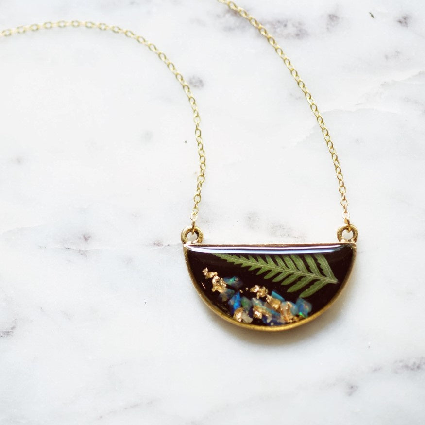 Gold plated necklace with a semicircle pendant featuring a small pressed fern frond and blue opal against a black resin background