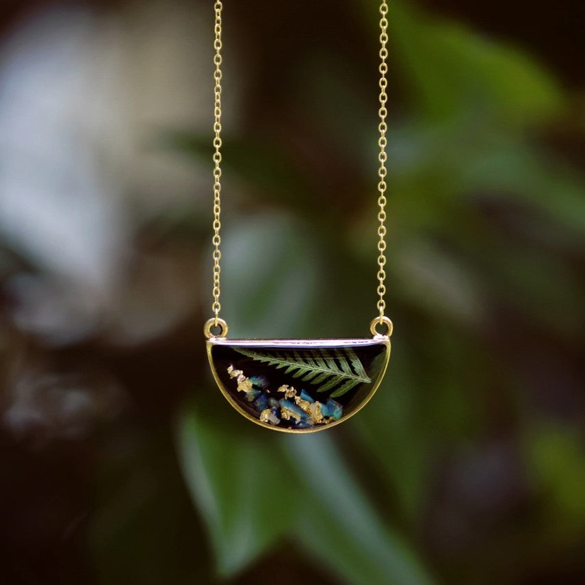 Gold plated necklace with a semicircle pendant featuring a small pressed fern frond and blue opal against a black resin background