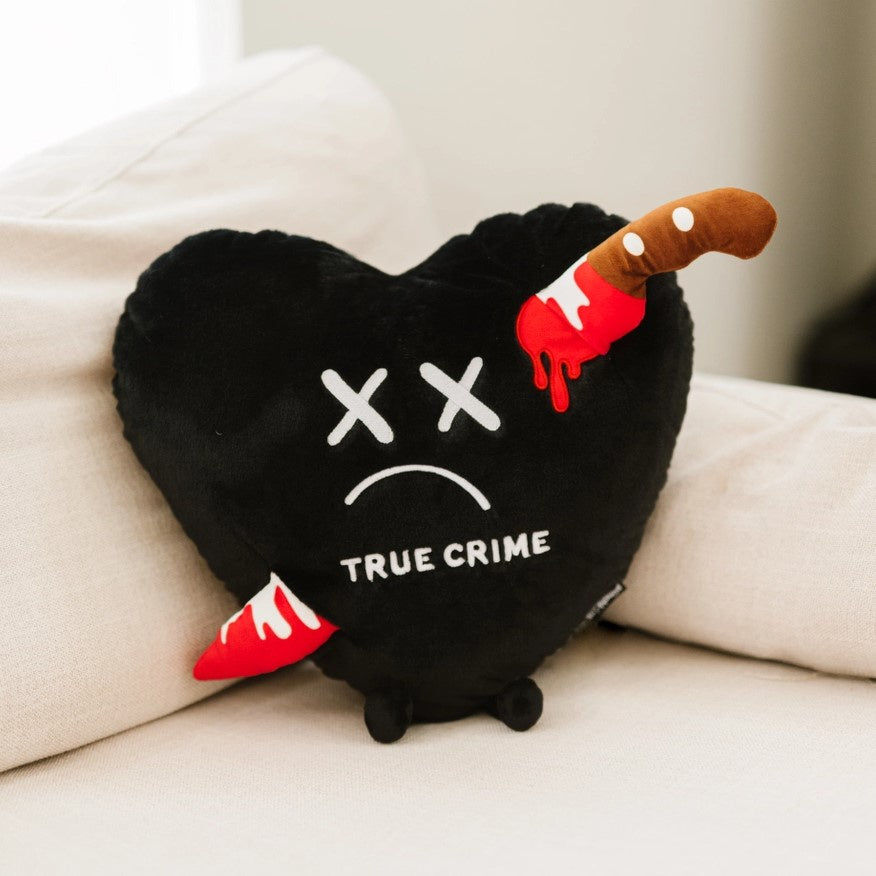 Large black heart plushie with x's for eyes and a bloody knife coming out either side. The words true crime are embroidered on it.