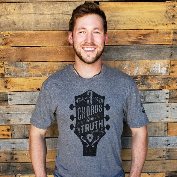 Man wearing a heathered gray t-shirt featuring a design of a guitar headstock with text "Three chords and the truth"