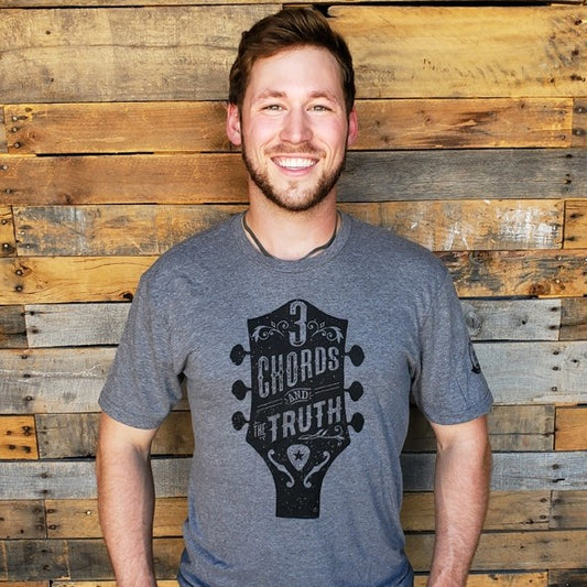 Man wearing a heathered gray t-shirt featuring a design of a guitar headstock with text "Three chords and the truth"