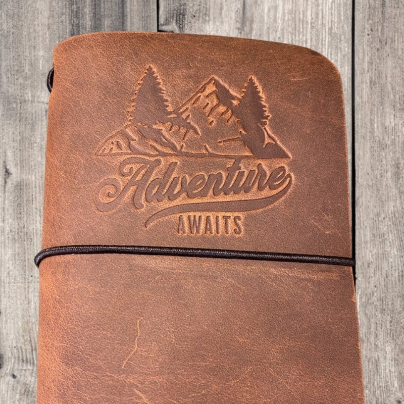 Leather journal with an image of a  mountain and the words "Adventure Awaits" engraved on the cover