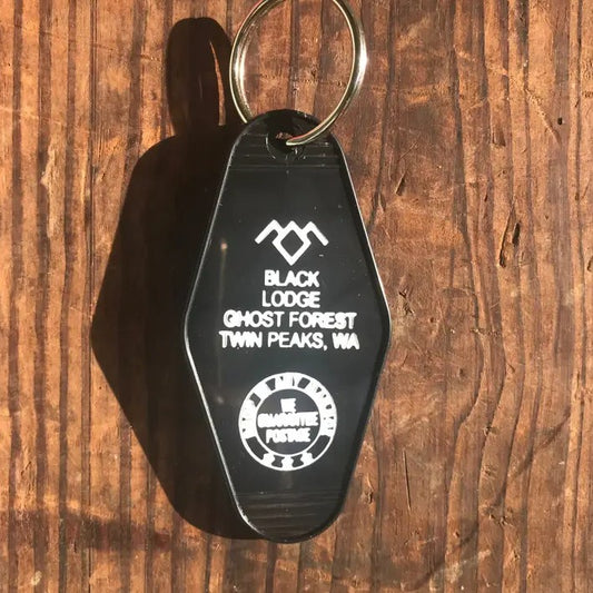 A black plastic keychain with a metal key ring, featuring white engraved text that reads 'BLACK LODGE GHOST FOREST TWIN PEAKS, WIA' and a circular badge, shown on a rustic wooden background.