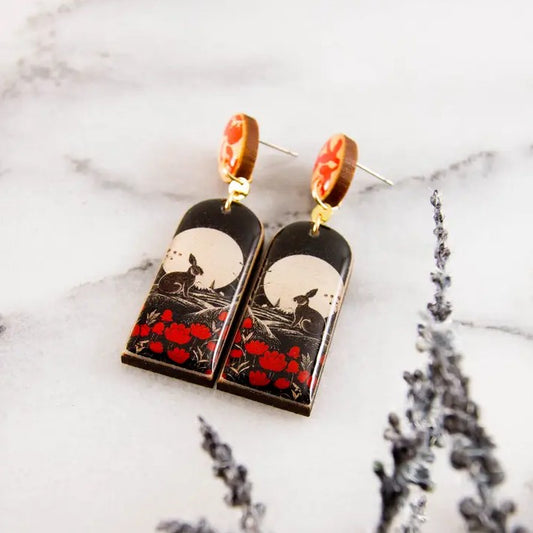 Pair of dangle earrings with a black, white and red image of a rabbit sitting in a poppy field 