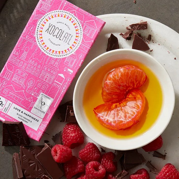 Chocolate bar with a pink wrapper next to a plate of dark chocolate, oranges and raspberries