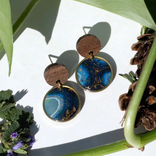 A pair of earrings with wooden studs and blue and gold resin charms hanging from them