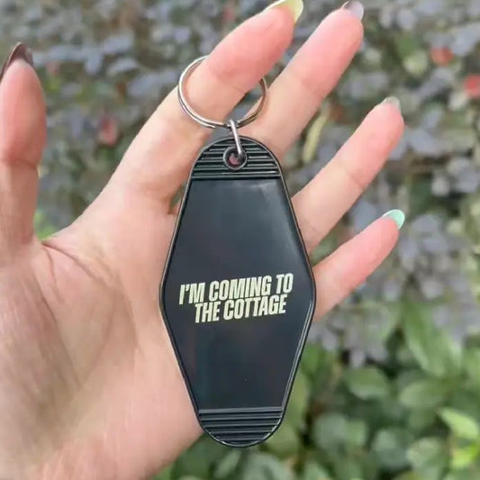 A hand holding a black plastic keychain with pale yellow bold text "I'M COMING TO THE COTTAGE"