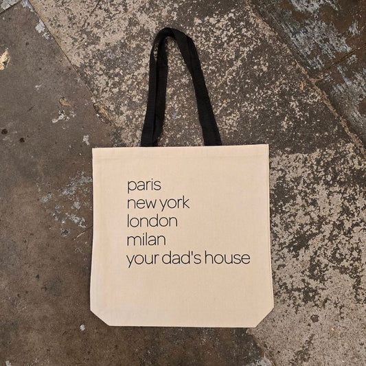 A beige canvas tote bag with black straps. The bag features black text "Paris, New York, London, Milan, Your Dad's House"