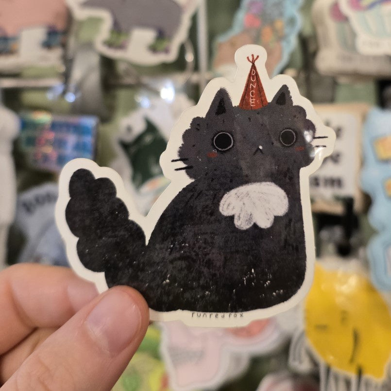 A small vinyl sticker featuring a cartoon black cat with large eyes wearing a tiny red dunce cap.