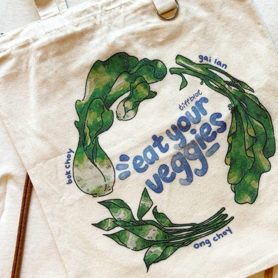 Beige tote bag with green leafy vegetables illustrated in watercolor style and blue text "eat your veggies"