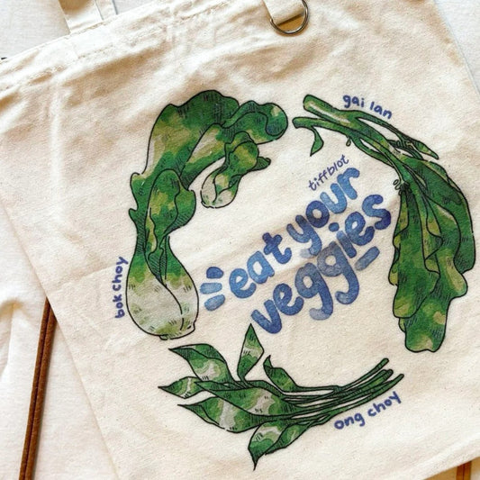 Beige tote bag with green leafy vegetables illustrated in watercolor style and blue text "eat your veggies"
