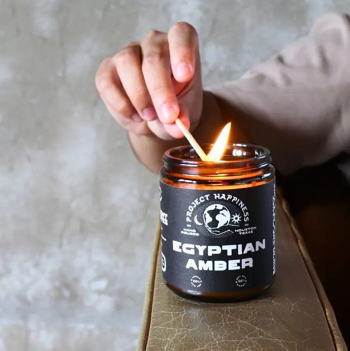 Hand lighting an Egyptian Amber candle with a match, against a neutral background.