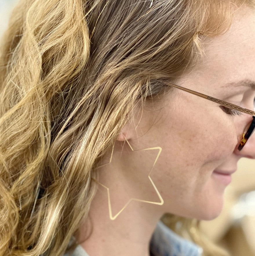 A person wearing 2.5 inch by 2.5 inch star shaped metal earrings with a satin finish 