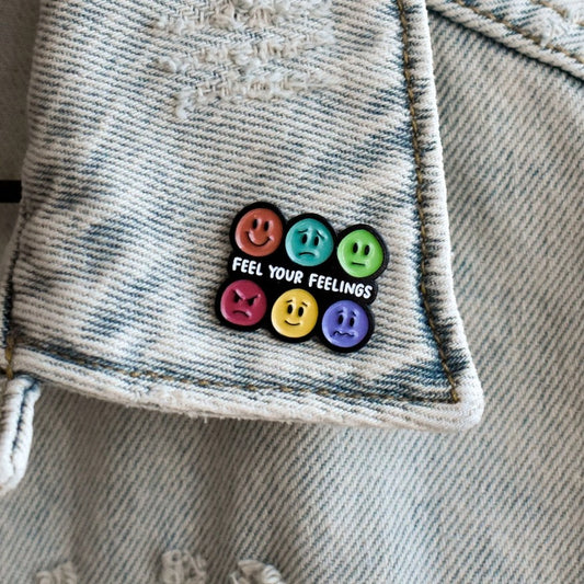 Enamel pin featuring rainbow color faces with different expressions and text that reads "Feel your feelings"