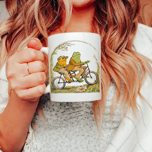 Person holding a white mug with an illustration of Frog and Toad riding a bicycle on it