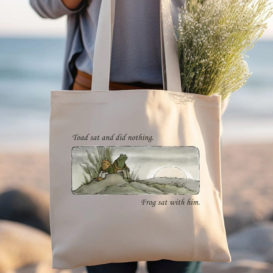 A tote bag featuring an image of frog and toad sitting next to each other on a grassy hill and captioned "Toad sat and did nothing. Frog sat with him."