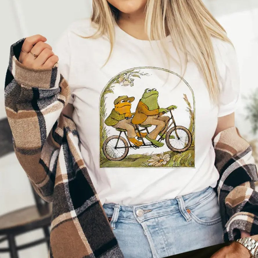 White t-shirt featuring an illustration of Frog and Toad riding a tandem bicycle